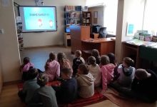 Dzieci siedzą na podłodze w czytelni i oglądają na tablicy multimedialnej prezentację