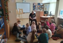 Dzieci siedzą na kocach w czytelni biblioteki i słuchają opowiadania czytanego przez nauczyciela