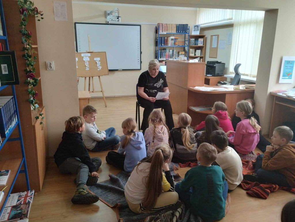 Dzieci siedzą na kocach w czytelni biblioteki i słuchają opowiadania czytanego przez nauczyciela