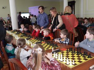 Dzieci siedzą przy stole z szachownicami. Za nimi stoją dorośli opiekunowie.